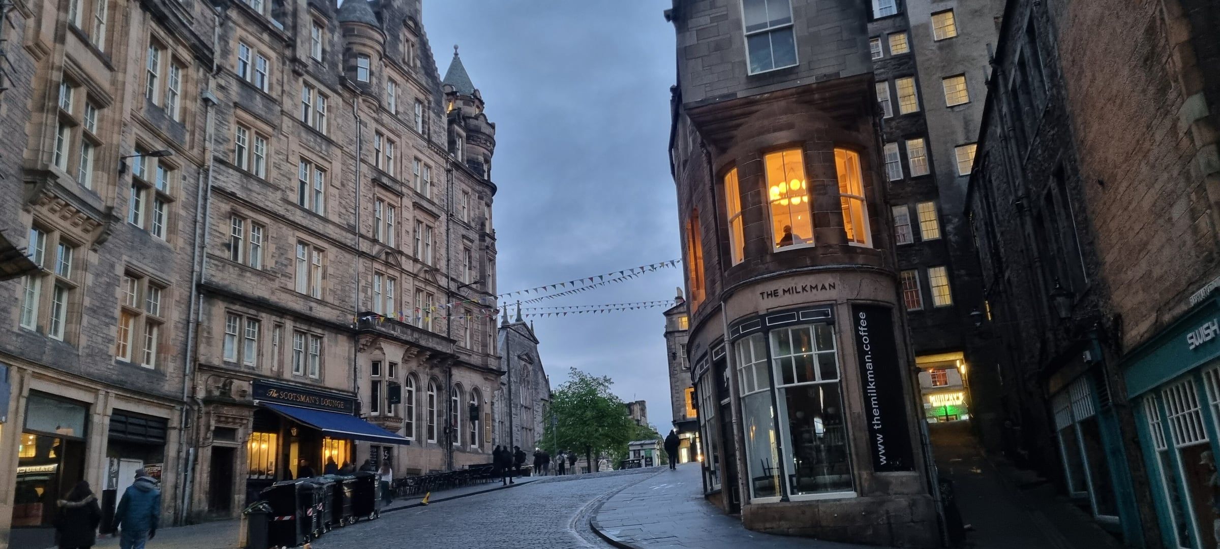 Narrow, curving Old Town street in low evening light, lined with stone tenements, lit shopfronts and a corner café.