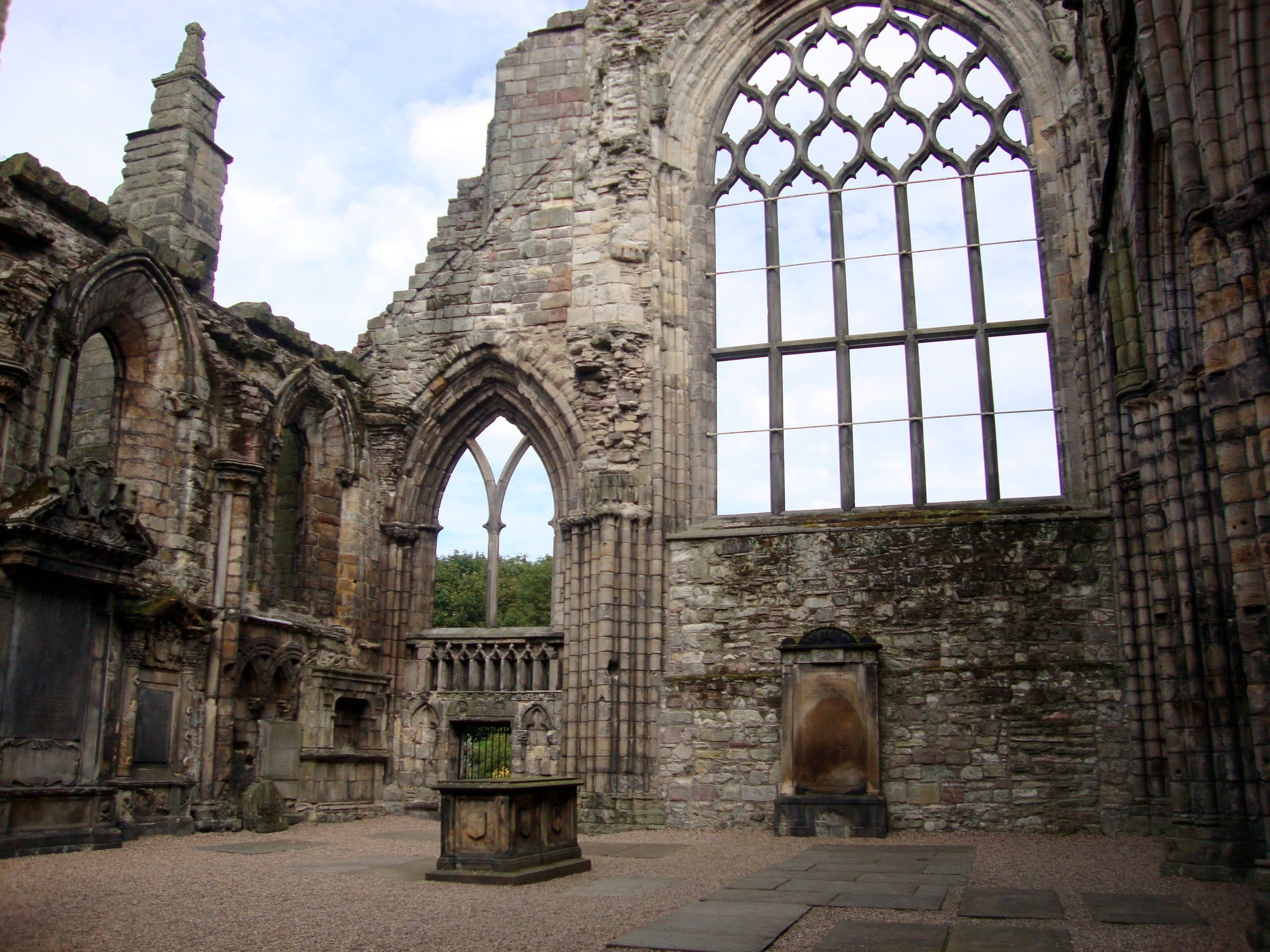 Roofless nave of Holyrood Abbey, with a tall Gothic window, pointed arches and a stone tomb in the foreground.