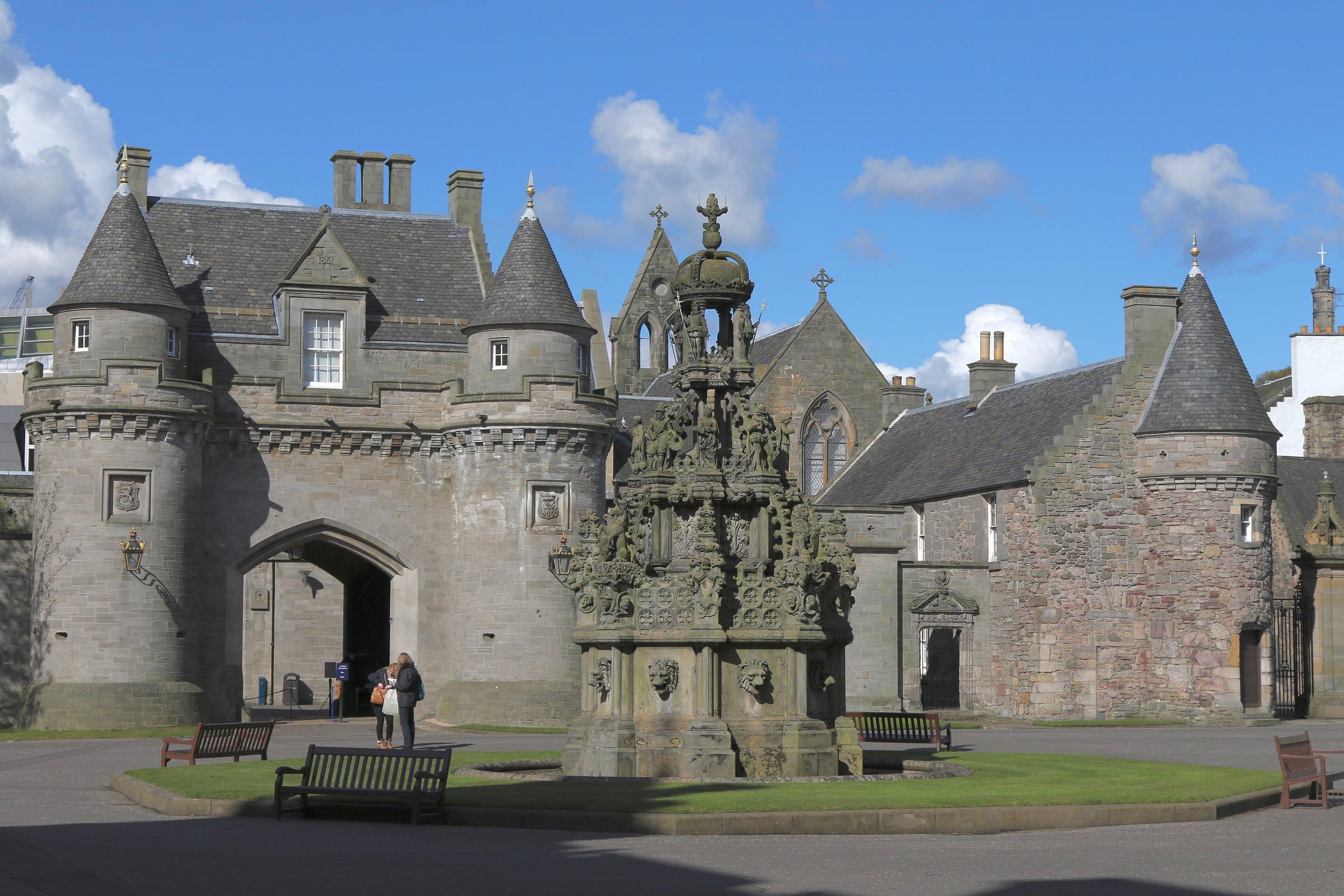 Stone gatehouse with turrets and an ornate carved fountain in the palace forecourt, under a partly cloudy sky.